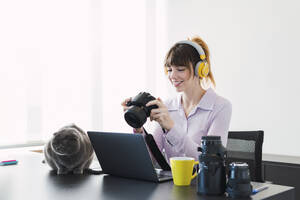 Glückliche Geschäftsfrau mit drahtlosen Kopfhörern, die mit der Kamera am Schreibtisch sitzt - PNAF05261