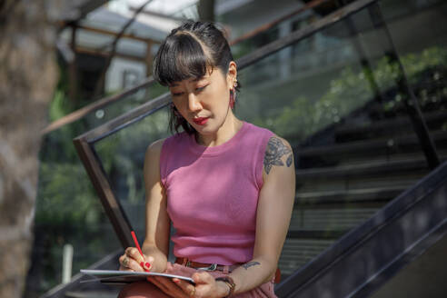Frau zeichnet mit Bleistift auf Skizzenblock sitzend vor einem Geländer - IKF00141