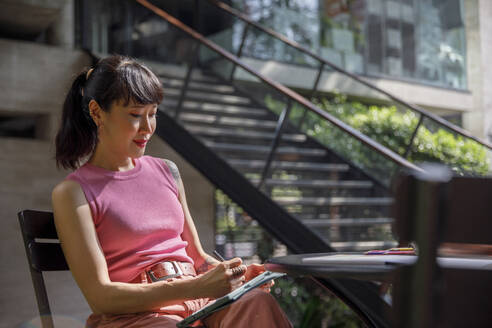 Woman using graphics tablet sitting on chair - IKF00146