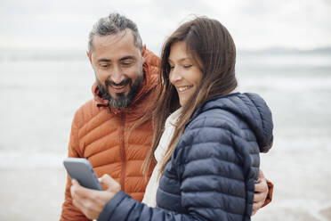 Happy man and woman using smart phone at beach - JOSEF18473