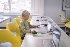 Blonder Junge mit Laptop auf einem Stuhl sitzend in der Schule - NJAF00336