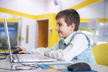 Smiling boy using laptop sitting at desk in school - NJAF00340