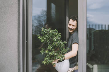 Glücklicher Mann mit Topfpflanze vor Glastür stehend - NDEF00535
