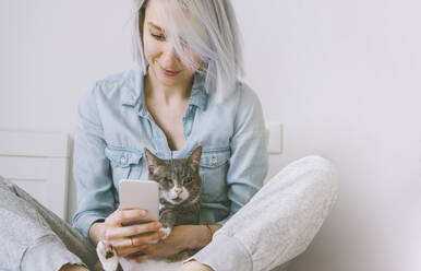 Smiling woman holding cat and using smart phone at home - NDEF00543