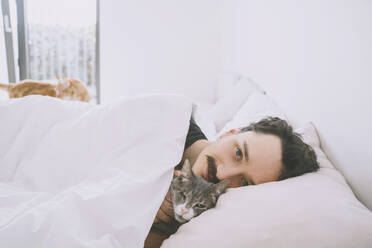 Lächelnder Mann entspannt sich auf dem Bett mit Katze zu Hause - NDEF00558