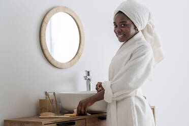 Smiling woman wearing bathrobe standing in bathroom at home - AAZF00305