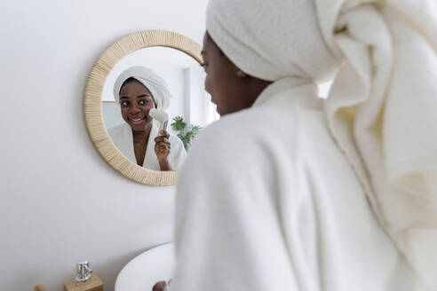 Lächelnde Frau mit Gesichtsmassagegerät im Spiegel zu Hause - AAZF00307