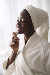 Smiling woman wearing bathrobe using face brush at home - AAZF00318