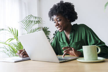 Happy businesswoman with laptop writing on note pad - AAZF00352