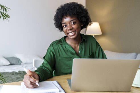 Glückliche Geschäftsfrau mit Stift und Laptop am Schreibtisch sitzend - AAZF00353