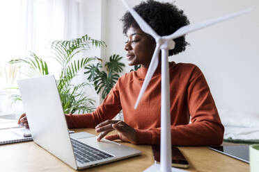 Smiling businesswoman sitting with laptop and wind turbine model at desk - AAZF00369