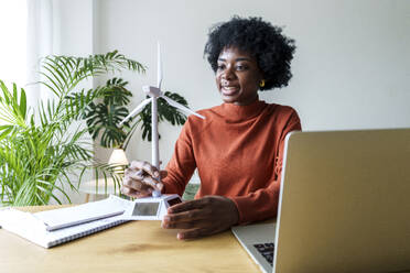 Smiling businesswoman with model wind turbine sitting at desk - AAZF00371