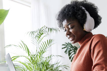 Happy young businesswoman wearing wireless headphones working on laptop - AAZF00382