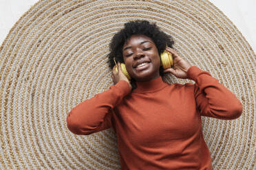 Glückliche junge Frau mit geschlossenen Augen, die drahtlose Kopfhörer trägt und auf einem Teppich liegt - AAZF00394