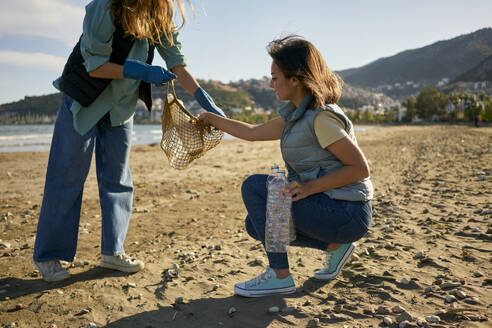 Woman putting plastic bottle in mesh bag held by volunteer at beach - ANNF00108