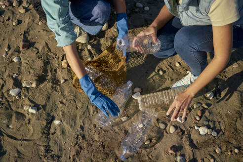 Women picking up plastic bottles amidst pebbles at beach on sunny day - ANNF00110