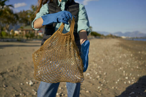 Volunteer holding mesh bag with plastic bottles at beach - ANNF00111