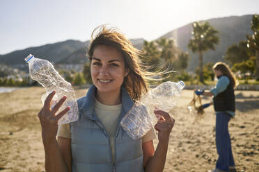 Smiling woman holding crumpled plastic bottles at beach - ANNF00112