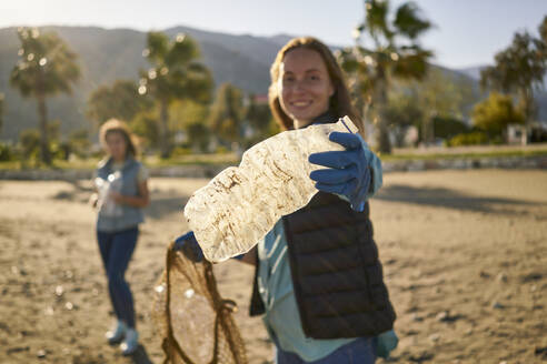 Lächelnde Aktivistin mit Plastikflasche am Strand an einem sonnigen Tag - ANNF00113