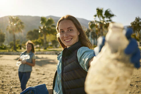 Happy environmentalist holding empty plastic bottle at beach - ANNF00115