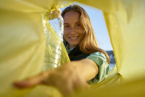 Smiling volunteer putting plastic bottles in yellow garbage bag - ANNF00121