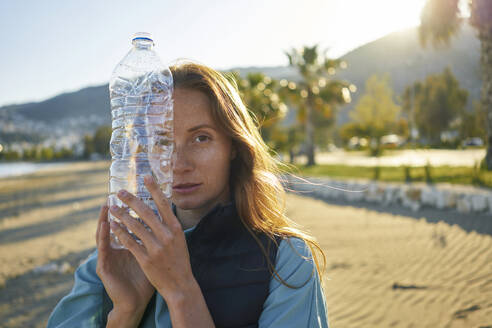 Volunteer holding empty plastic bottle near face at beach - ANNF00125