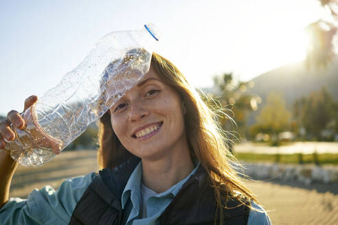 Smiling volunteer holding plastic bottle near forehead at beach - ANNF00126