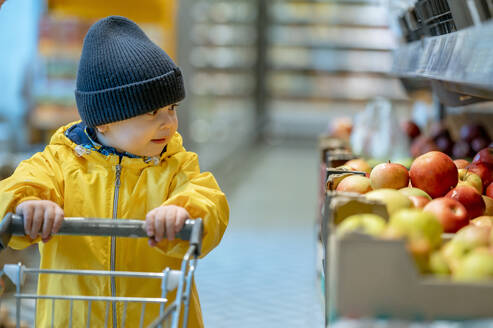 Junge sieht sich Äpfel an und hält einen Einkaufswagen im Geschäft - ANAF01241