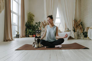 Lächelnde Frau, die mit ihrem Schnauzer-Hund auf einer Übungsmatte zu Hause Yoga übt - YTF00736