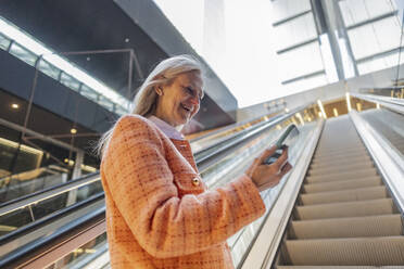 Lächelnde Geschäftsfrau mit Smartphone auf der Rolltreppe stehend - JCCMF10316