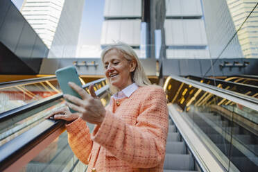 Smiling businesswoman using mobile phone standing on escalator - JCCMF10317