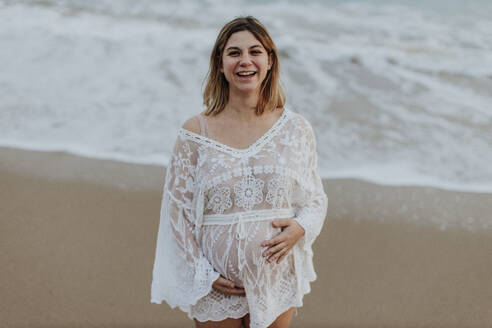 Glückliche schwangere Frau am Strand stehend - GMLF01396