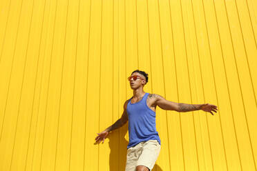 Young man with arms outstretched dancing in front of yellow wall on sunny day - SYEF00338