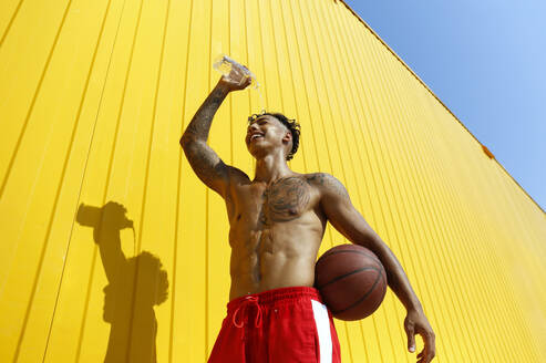 Smiling man pouring water on head standing with basketball near yellow wall - SYEF00340