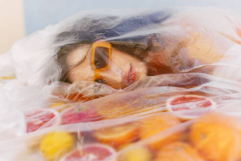 Woman wearing sunglasses by citrus fruits covered in plastic foil - EGHF00766