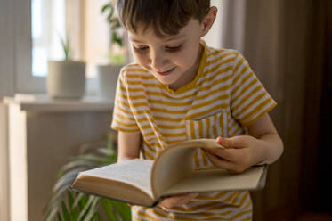 Smiling boy flipping pages of book at home - ANAF01247