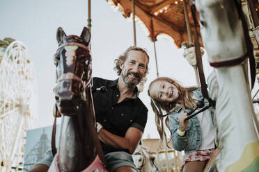 Father and daughter enjoying carousel ride at amusement park - GMLF01411