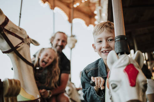 Smiling boy enjoying carousel ride with family at amusement park - GMLF01414