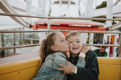 Girl kissing brother sitting in ferris wheel at amusement park - GMLF01415