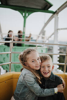 Lächelndes Mädchen, das seinen Bruder umarmt, der im Riesenrad im Vergnügungspark sitzt - GMLF01419