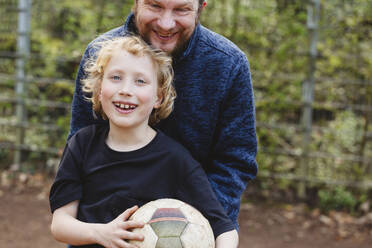 Father embracing happy son holding soccer ball - IHF01336