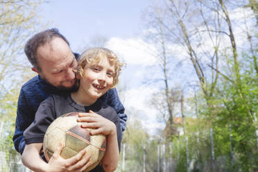 Playful father embracing smiling son holding soccer ball - IHF01337