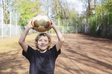 Lächelnder Junge hält Fußball auf dem Spielplatz - IHF01338
