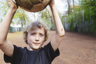 Blonder Junge mit Bluterguss am Unterarm hält Fußball auf dem Spielplatz - IHF01339