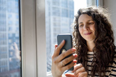 Smiling woman using smart phone near window - ANAF01249
