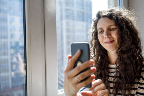 Lächelnde Frau mit Smartphone am Fenster - ANAF01249