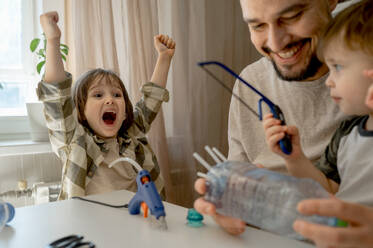 Fröhlicher Junge mit Vater und Bruder bei der Herstellung von Plastikflaschen für Autos zu Hause - ANAF01259