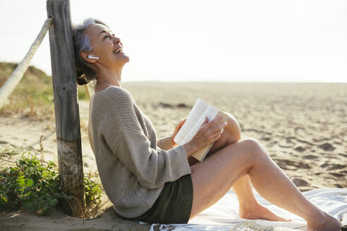 Glückliche reife Frau mit Buch am Strand sitzend - EBSF03212