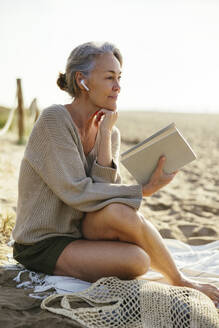 Nachdenkliche Frau mit Hand am Kinn und Buch am Strand - EBSF03214