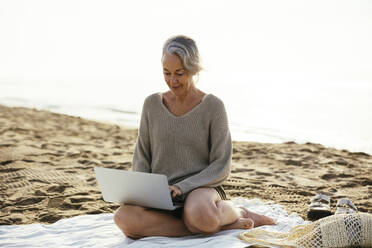 Mature woman using laptop sitting at beach - EBSF03221